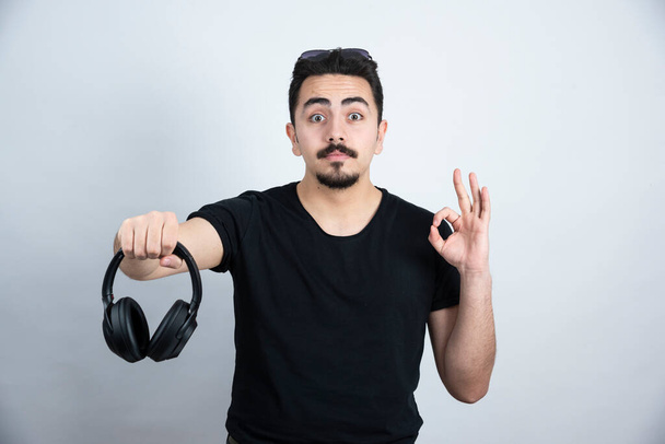 Brunette man model holding headphones and showing ok gesture against white wall. High quality photo - Foto, Imagen
