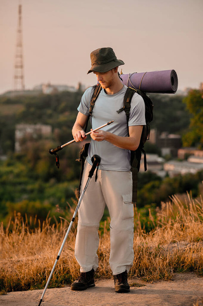 Ένας νεαρός άνδρας με σακίδιο στέκεται στο λόφο με τα πόδια μπαστούνια στα χέρια - Φωτογραφία, εικόνα