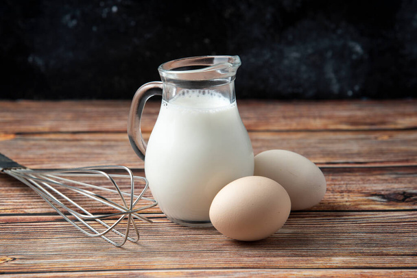 Glass jug of milk, eggs and whisker on wooden table. High quality photo - Fotoğraf, Görsel