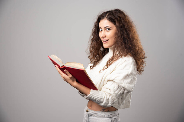 Fiatal, mosolygós nő, vörös könyvvel a szürke falon. Kiváló minőségű fénykép - Fotó, kép