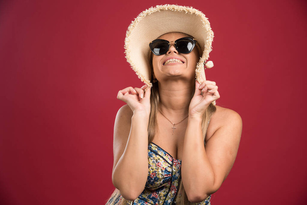 Ξανθιά γυναίκα με καπέλο και γυαλιά να γελάει σε κόκκινο φόντο. Υψηλής ποιότητας φωτογραφία - Φωτογραφία, εικόνα