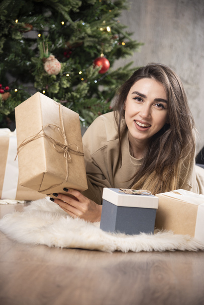 Χαμογελαστή γυναίκα ξαπλωμένη στο χνουδωτό χαλί και δείχνοντας ένα χριστουγεννιάτικο δώρο. Υψηλής ποιότητας φωτογραφία - Φωτογραφία, εικόνα