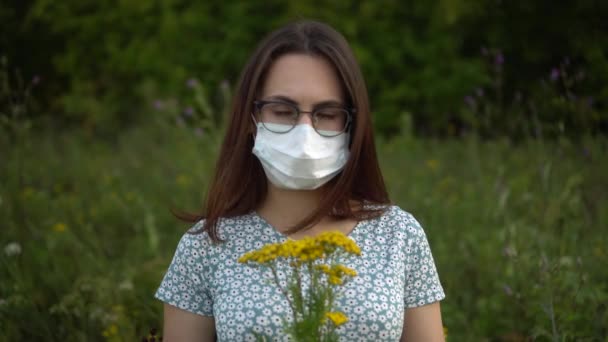 Eine junge Frau in medizinischer Maske riecht Blumen und schüttelt den Kopf, weil sie nicht riecht. Mädchen mit Brille und Kleid in der Natur. - Filmmaterial, Video