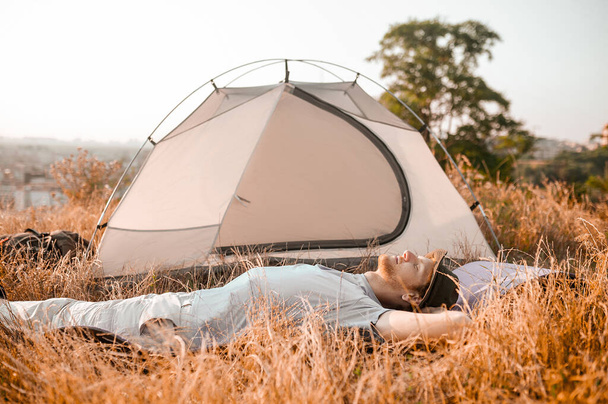 Een man die op het gras bij zijn tent ligt en ontspannen kijkt. - Foto, afbeelding