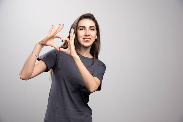 Junge Frau in grauem T-Shirt vor grauem Hintergrund, die mit den Händen Herzsymbole in Form bringt. Hochwertiges Foto - Foto, Bild