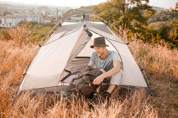 A man in a hat packing his backpack while sitting near the tent - Photo, Image