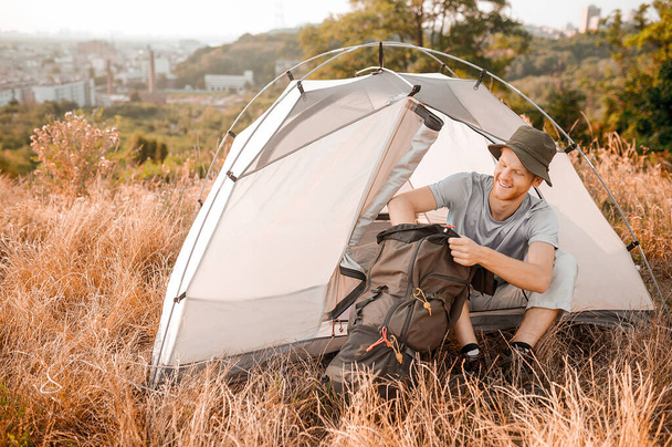 Un uomo con un cappello che impacchetta lo zaino mentre è seduto vicino alla tenda - Foto, immagini