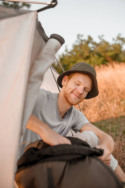Uśmiechnięty mężczyzna wygląda na zadowolonego siedząc blisko namiotu. - Zdjęcie, obraz