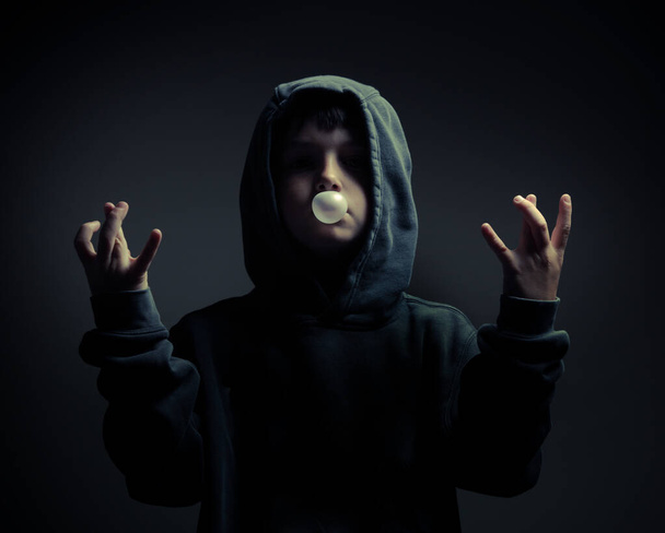 Un retrato de un niño en la sudadera con capucha, soplando el chicle, en el ambiente oscuro - Foto, Imagen
