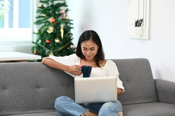 Μια χαρούμενη γυναίκα ξοδεύει το Σαββατοκύριακο στο σπίτι πίνοντας καφέ και χρησιμοποιώντας φορητό υπολογιστή στον καναπέ. - Φωτογραφία, εικόνα