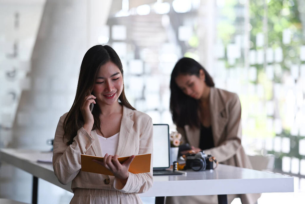 A beautiful businesswoman talking on mobile phone and reading information in notebook at office. - Foto, imagen