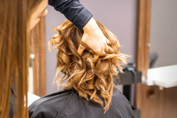 Female hairdresser checks brown curly hairstyle of a young caucasian woman in beauty salon - Photo, Image