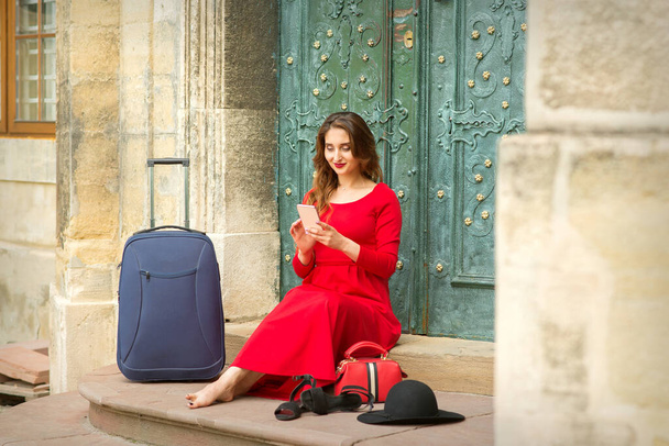 Mooie jonge blanke vrouw zittend op de trap bij de deur met reiskoffer en smartphone in lange rode jurk - Foto, afbeelding