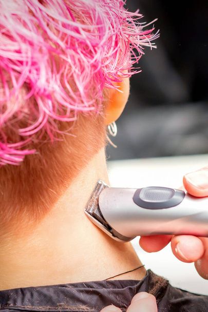 Vista posterior de la mano de peluquero afeitado nuca y cuello con trimmer eléctrico de mujer joven caucásica con pelo corto de color rosa en el salón de belleza - Foto, imagen