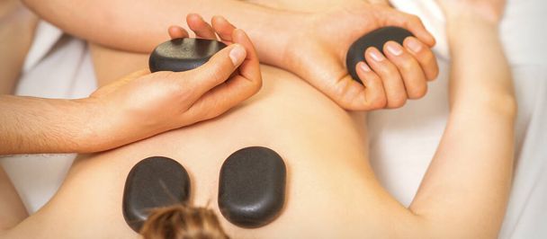 Массаж горячими камнями на женской спине с руками массажиста, держащего черные массажные камни в спа-салоне - Фото, изображение