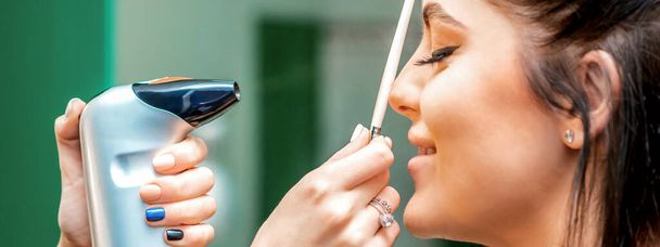 Seitenansicht eines Make-up-Artist mit Hilfe eines Aerographen, der ein Airbrush Make-up-Fundament auf einem weiblichen Gesicht in einem Schönheitssalon erstellt - Foto, Bild