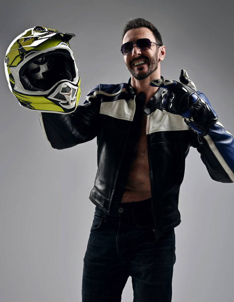Laughing man biker in sunglasses, jeans and motorcycle jacket on naked body stands pointing finger at helmet he holds - Photo, image