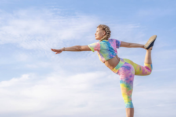 Молодая красивая блондинка в спортивном костюме стоит на одной ноге на фоне голубого летнего неба и сохраняет равновесие. Спортивная гимнастика на открытом воздухе. - Фото, изображение