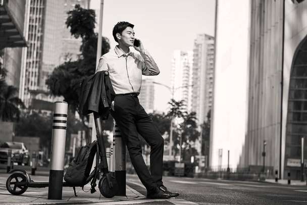 young asian businessman standing next to his electric scooter on street making a call using cellphone in downtow of modern city, black and white - Photo, Image