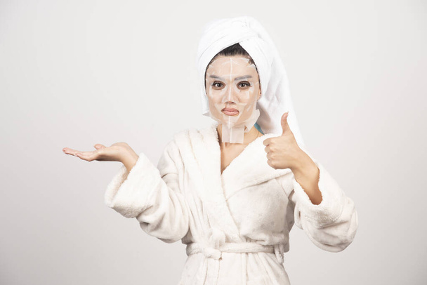 Nainen näyttää peukalo pystyssä ja yllään kylpytakki ja pyyhe kasvot. Laadukas kuva - Valokuva, kuva