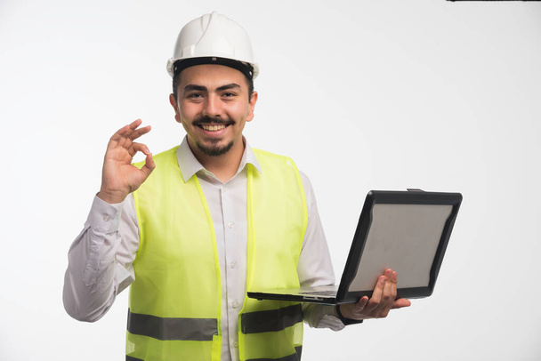 Ingeniero en uniforme sosteniendo un ordenador portátil y haciendo señal positiva. Foto de alta calidad - Foto, Imagen