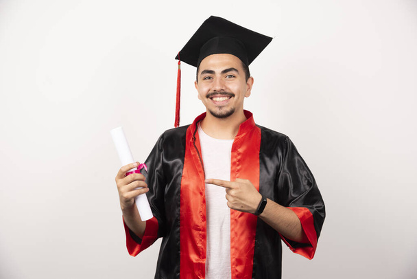 Счастливый студент указывает на диплом на белом фоне. Высокое качество фото - Фото, изображение