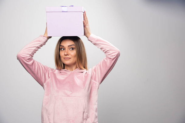 Ξανθιά κυρία με ροζ μπλούζα κρατά ένα κουτί δώρου πάνω από το κεφάλι της και κουνιέται. Υψηλής ποιότητας φωτογραφία - Φωτογραφία, εικόνα