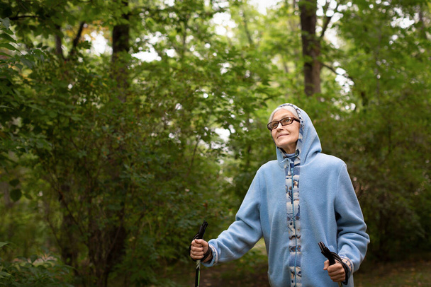 Waist up portrait of elderly woman. Caucasian old lady in blue hoodie and glasses smiling and does nordic walking in the park outdoor in late summer, selective focus. - Фото, зображення
