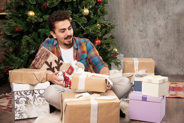 Lächelnder gutaussehender Mann sitzt mit Weihnachtsgeschenken und wählt das beste. Hochwertiges Foto - Foto, Bild