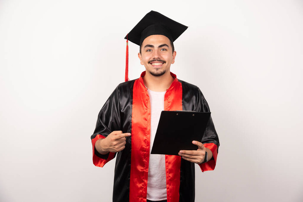 Junger Doktorand zeigt auf sein Diplom auf weißem Hintergrund. Hochwertiges Foto - Foto, Bild