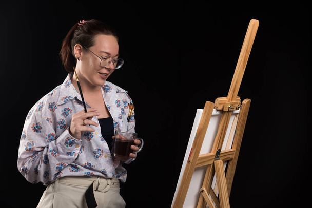 Donna artista tenendo pennello e guardando tela su sfondo nero  - Foto, immagini