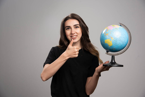 Happy woman with globe making thumbs up on gray background. High quality photo - Foto, Bild
