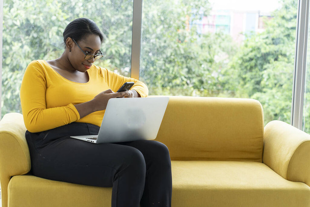 Afroamerikanerin sitzt auf gelbem Sofa, tippt Nachrichten auf dem Smartphone und arbeitet von zu Hause aus am Laptop im Wohnzimmer.  - Foto, Bild