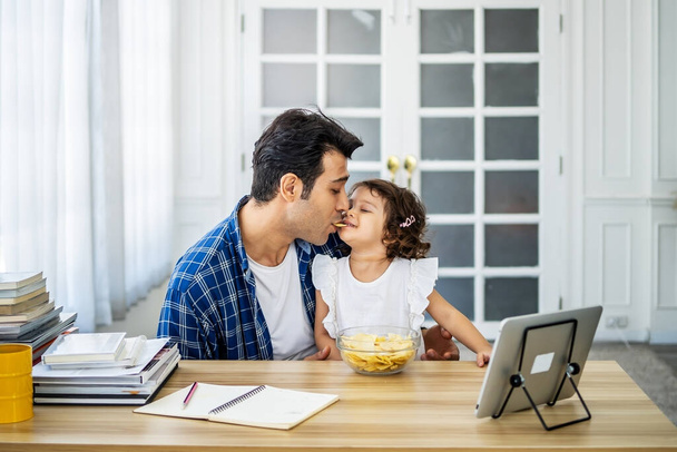 Caucasian family young father eating potato chips in mounth with little daughter together and watch cartoons educational online program - 写真・画像