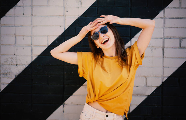Portrait of a young happy girl woman in sunglasses and a yellow t-shirt on the street a wall background - Фото, зображення