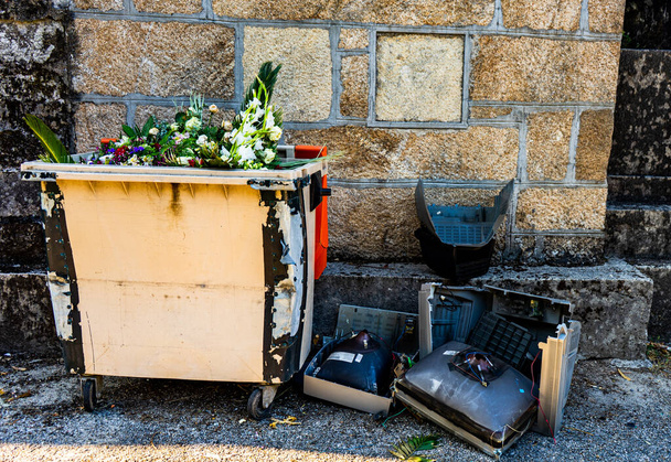 Müllcontainer neben einer Steinmauer voller Blumen und mit Bildschirmen alter Fernseher auf dem Boden - Foto, Bild