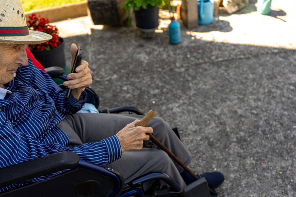 ηλικιωμένη γυναίκα με ψάθινο καπέλο παρακολουθεί κινητό τηλέφωνο από την αναπηρική της καρέκλα - Φωτογραφία, εικόνα