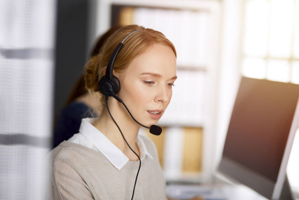 Friendly caucasian businesswoman talking by headset in sunny office. Call center and diverse people group in business - Foto, Imagen