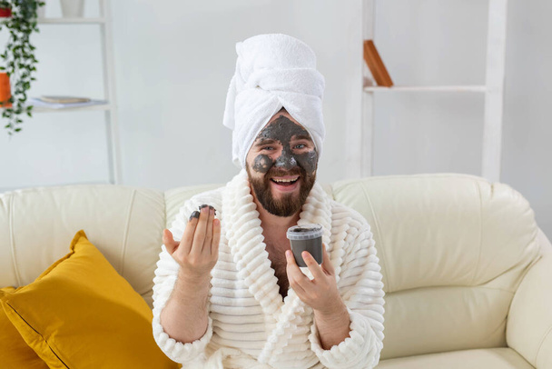 Zabawny brodaty mężczyzna bawiący się maską kosmetyczną na twarzy zrobioną z czarnej gliny. Mężczyźni pielęgnacja skóry, humor i spa w domu koncepcja - Zdjęcie, obraz