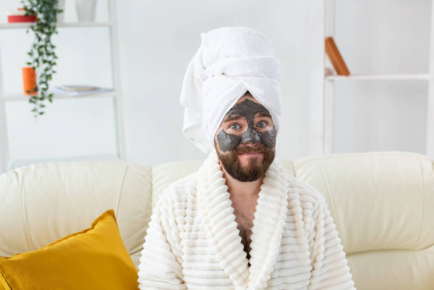 Brodaty, zabawny facet z maską kosmetyczną na twarzy zrobioną z czarnej gliny. Mężczyźni pielęgnacja skóry, humor i spa w domu koncepcja - Zdjęcie, obraz