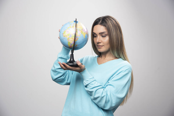 Mavi kazaklı sarışın kız elinde bir küre tutuyor ve dünya haritasını dikkatlice kontrol ediyor. Yüksek kalite fotoğraf - Fotoğraf, Görsel