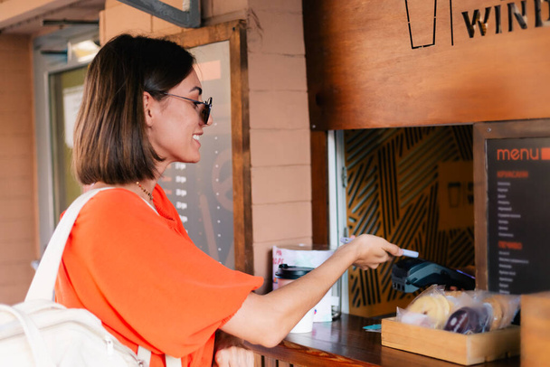 Vrouw koopt koffie buiten bij koffie venster met behulp van mobiele telefoon om te betalen - Foto, afbeelding