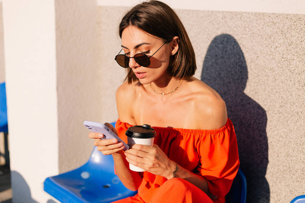 Tyylikäs nainen oransseissa vaatteissa auringonlaskun aikaan pyöräilyradan stadionilla kupillisella kahvia ja matkapuhelinta katso näyttöä - Valokuva, kuva