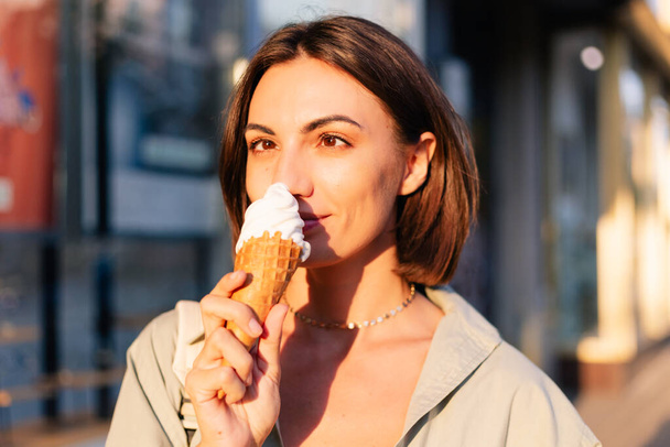 Yaz günbatımında kadın dışarıda dondurma yiyor. Şehir caddesinde neşeli, neşeli bir dondurma yiyor. - Fotoğraf, Görsel