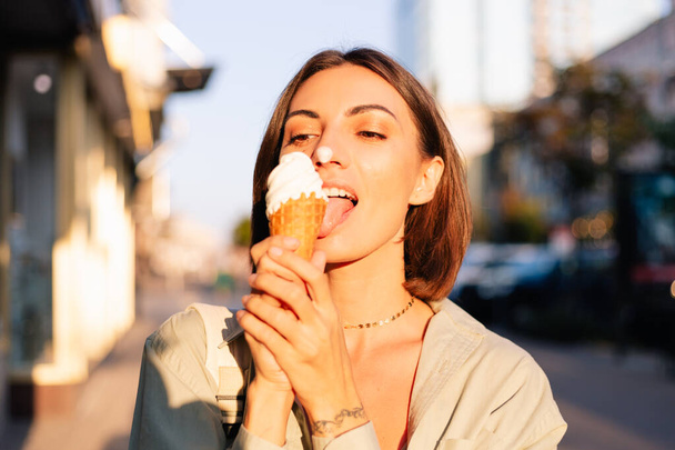 Femme en plein air à l'heure du coucher du soleil d'été ayant cône de crème glacée positif heureux ludique à la rue de la ville - Photo, image