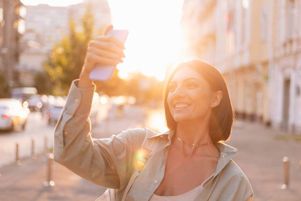Woman outdoor  at summer sunset time with mobile phone happy positive smile take photo video selfie - Photo, Image