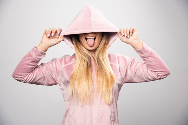 ピンクのスウェットシャツを着たブロンドの女性は、彼女の頭にパーカーを着用することによって喜びと肯定的なポーズを作ります。高品質の写真 - 写真・画像