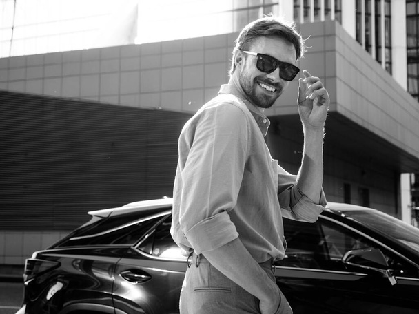 Portrait of handsome confident stylish hipster lambersexual model.Modern man dressed in shirt. Fashion male posing in the street background near skyscrapers in sunglasses. Near luxury car - Foto, immagini