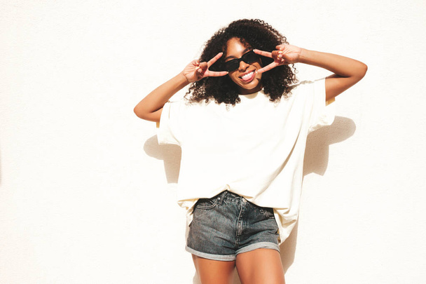 Beautiful black woman with afro curls hairstyle.Smiling hipster model in white t-shirt. Sexy carefree female posing in the street near white wall in sunglasses. Cheerful and happy.Shaws peace sign - Fotoğraf, Görsel