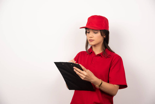 Giovane donna di consegna in uniforme rossa che scrive negli appunti su sfondo bianco. Foto di alta qualità - Foto, immagini